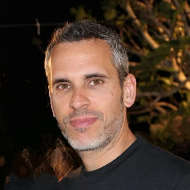 A man with short, salt-and-pepper hair and a trimmed beard is looking at the camera, standing outdoors at night with blurred trees in the background. He is wearing a dark shirt and has a neutral expression.